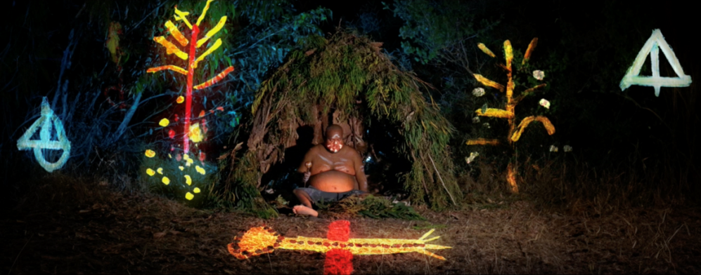 Patrick Carter artwork still, sitting before grass hut with neon lights around