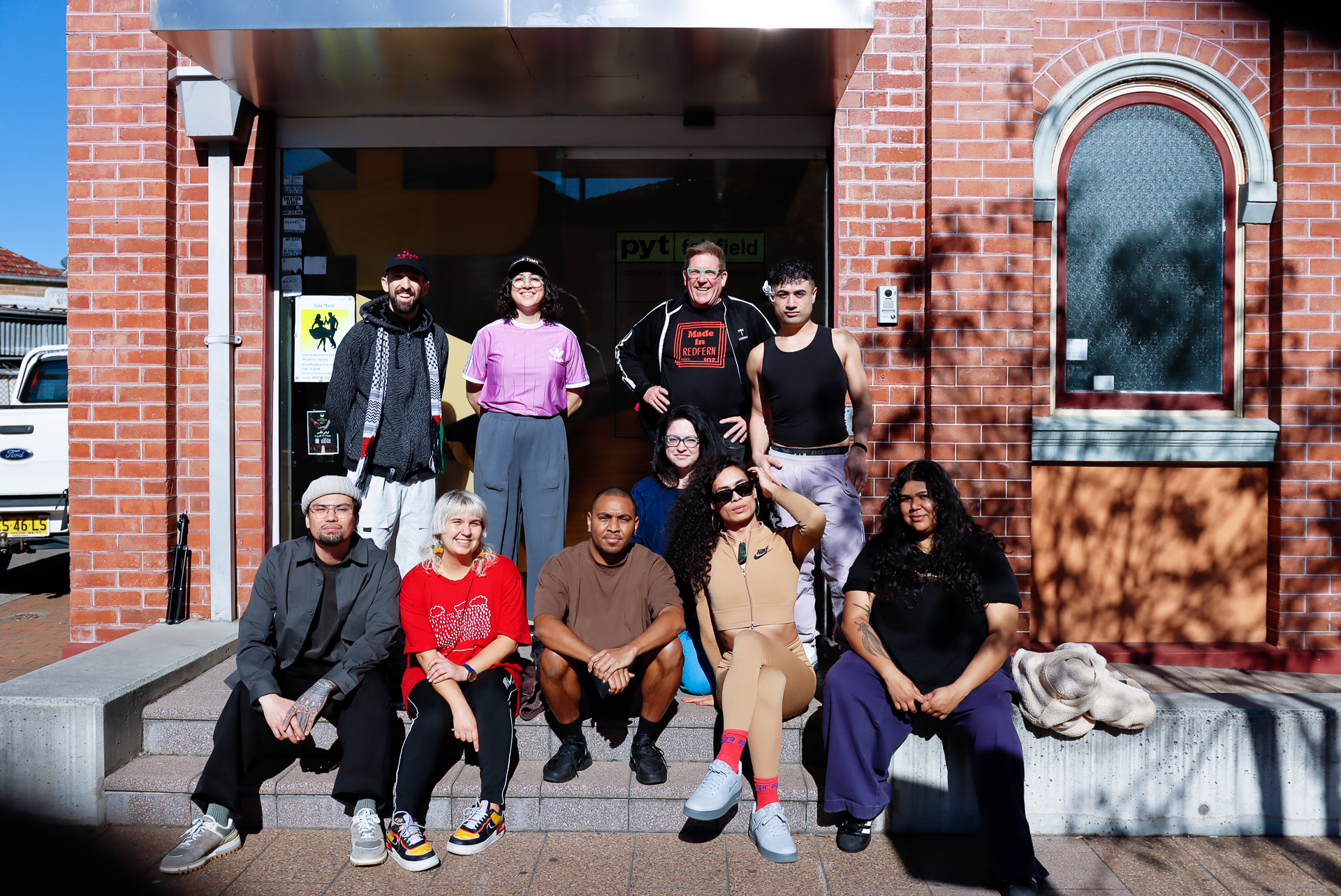 A photo of nine artists sitting on the front steps of a red building looking at the camera.