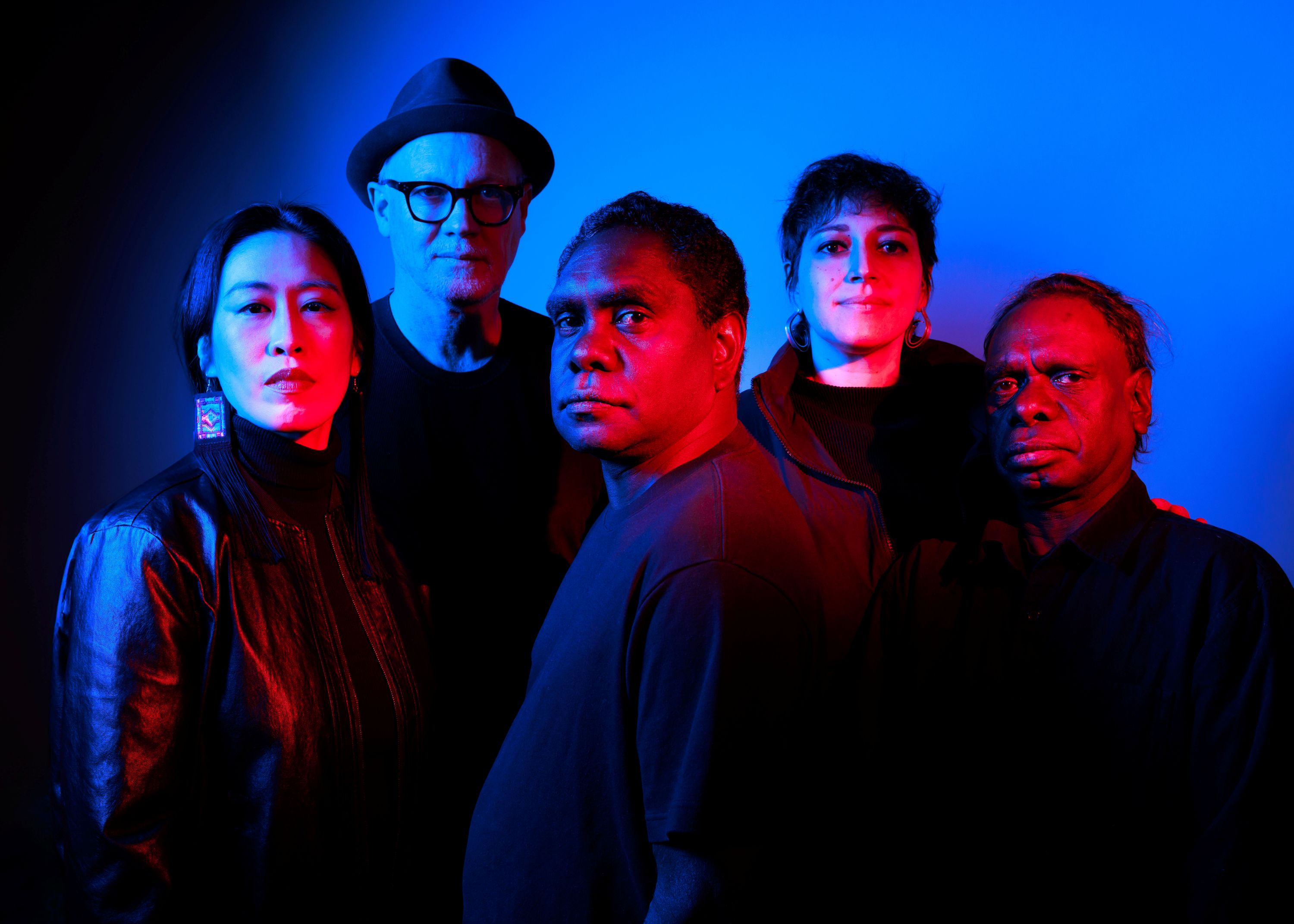 A photo of a group of 5 performers dressed all in black, looking at the camera as they are covered in dramatic red light, in front of a blue wall.
