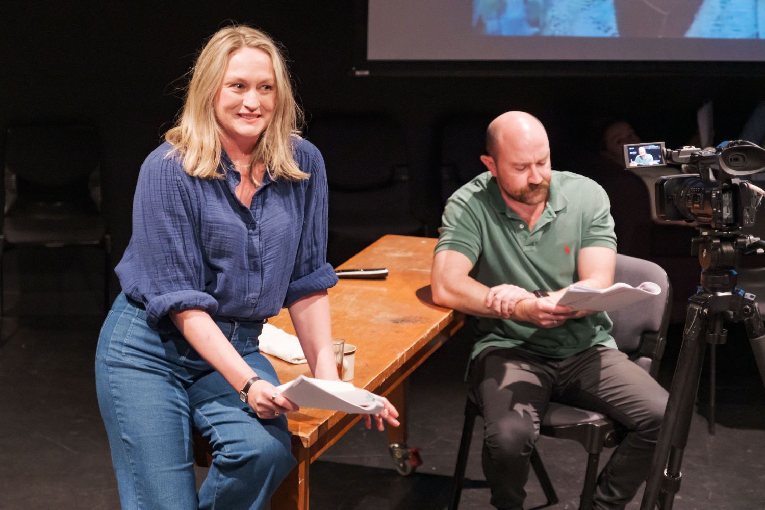 A photo of two performers rehearsing. One sits on a table holding a script but looking off camera, another sits at a chair, reading their script.