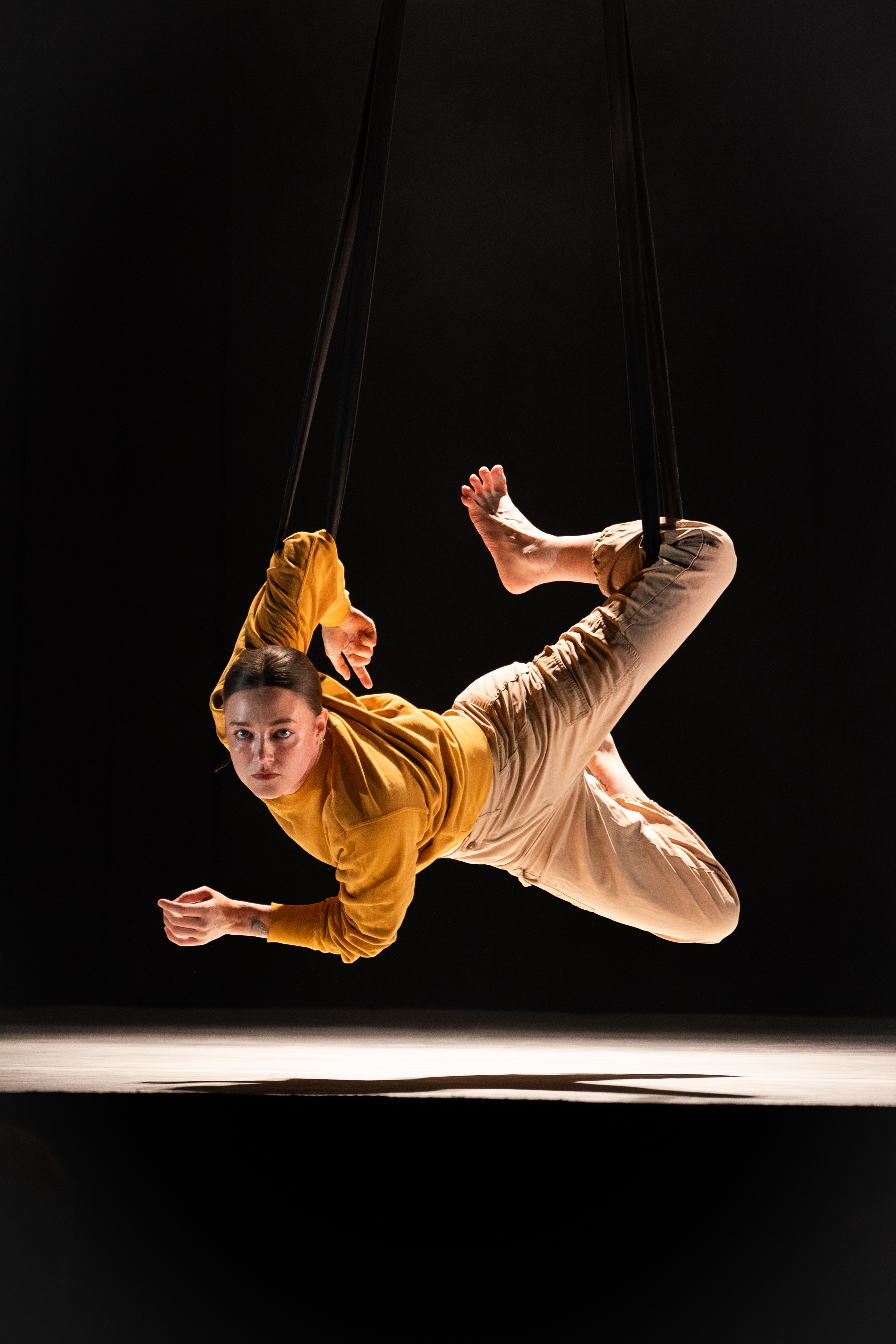 A circus performer hovers in the air about a foot away from the stage. They are contorting and looks straight at the camera.
