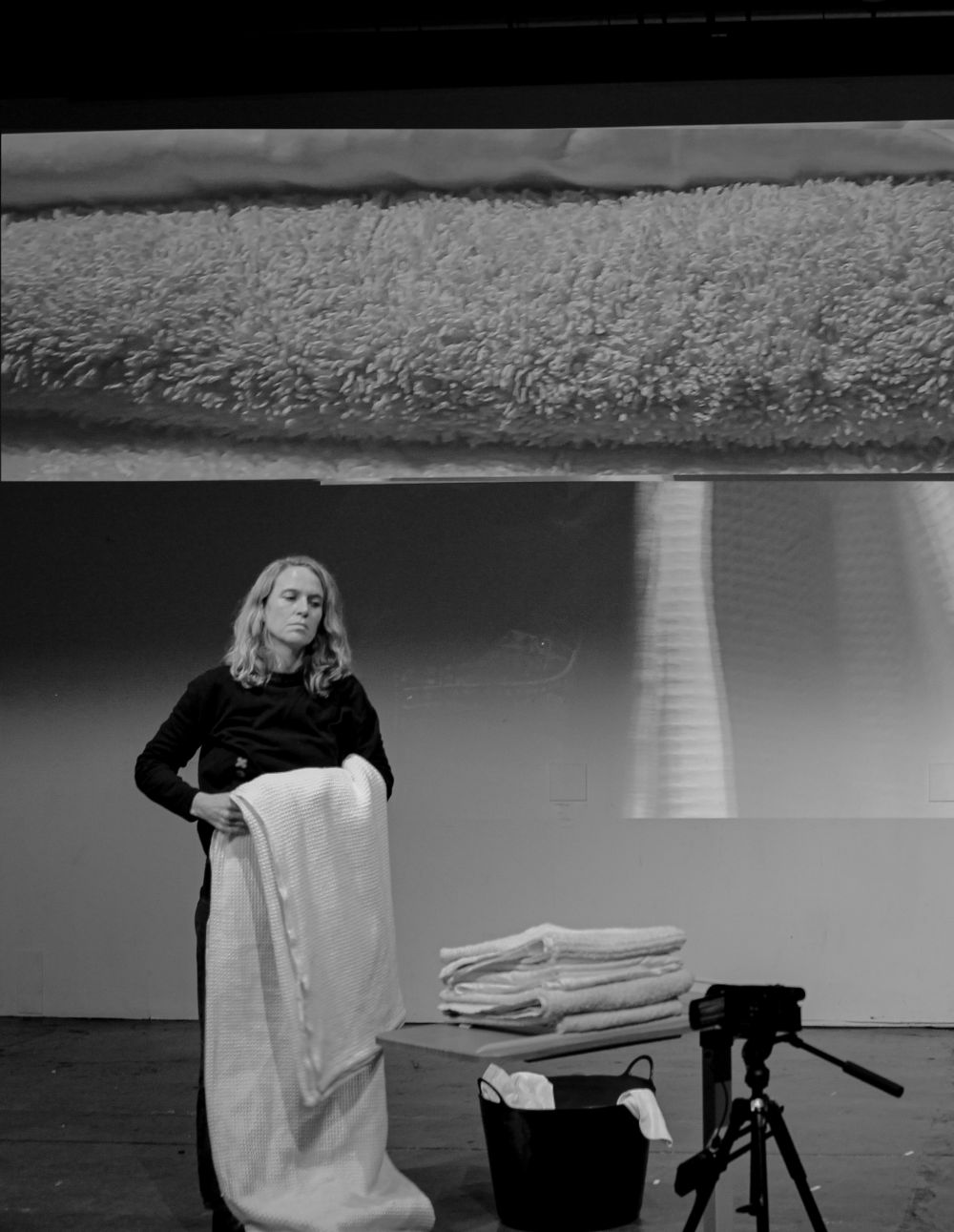 A middle aged woman stands looking blankly. She is folding a bed sheet. In front of her is a pile of folded towels and sheets with a camera pointing at them. Projected onto a wall behind her, is a close up projection of towels. The image is black and white. 