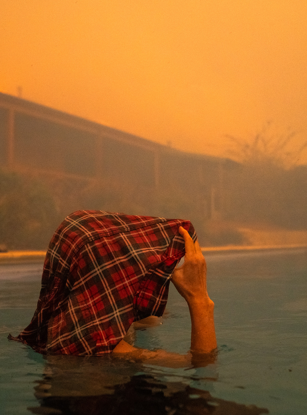 A person stands shoulder deep in a swimming pool holding a wet red, black and white checked flannel shirt over their head. Their face is not visible as they look to the right of the image. The house in the background is out of focus, the sky is brown and air is smoky.