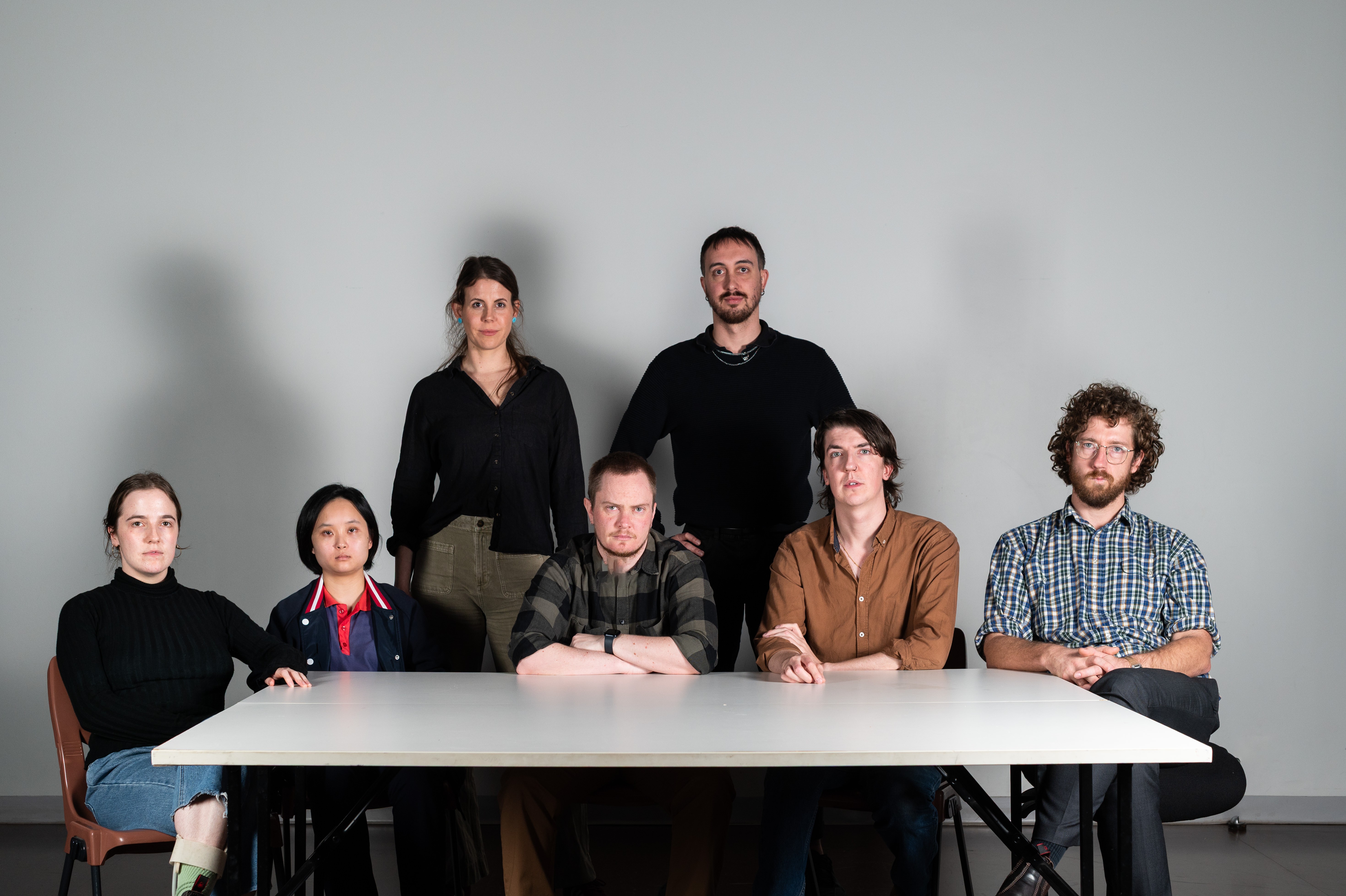The Milan Conference artists sit around a white table, dramatically lit and staring blankly at the camera.