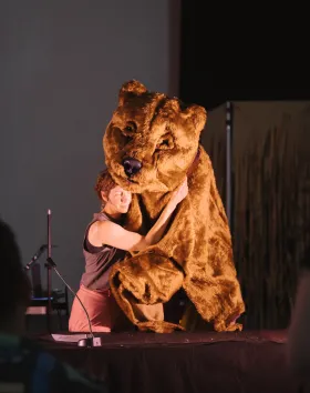 A photo of a performer and a large bear puppet on stage. They are embraced in a hug.