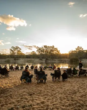 A photo of a riverbank at sunset. A group of people sit on the bank in foldout chairs.