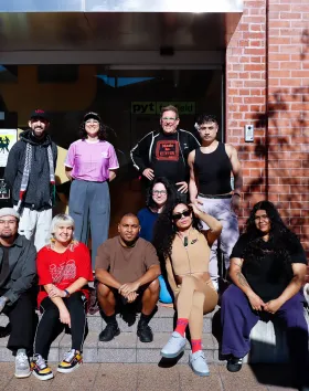 A photo of nine artists sitting on the front steps of a red building looking at the camera.