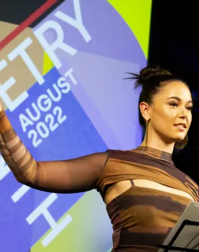 A photo of a performer standing on stage infront of a micrphone and music stand with one arm out the side as a dramatic gesture. Behind them a screen says Monthly Poetry.