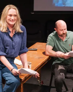 A photo of two performers rehearsing. One sits on a table holding a script but looking off camera, another sits at a chair, reading their script.