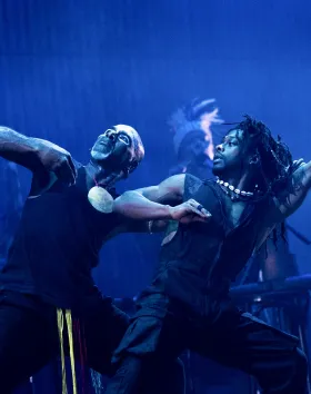 A photo of two dancers lunging side by side on stage surrounded by blue light.