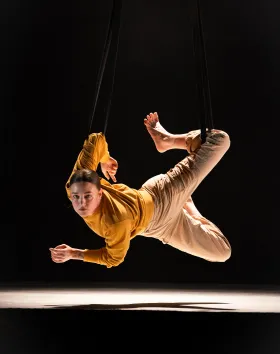 A circus performer hovers in the air about a foot away from the stage. They are contorting and looks straight at the camera.