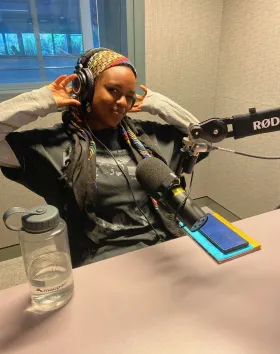 A photo of Rataj Abdullah in a radio studio smiling at the camera in front of a microphone.