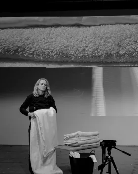 A middle aged woman stands looking blankly. She is folding a bed sheet. In front of her is a pile of folded towels and sheets with a camera pointing at them. Projected onto a wall behind her, is a close up projection of towels. The image is black and white. 