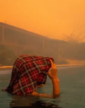 A person stands shoulder deep in a swimming pool holding a wet red, black and white checked flannel shirt over their head. Their face is not visible as they look to the right of the image. The house in the background is out of focus, the sky is brown and air is smoky.