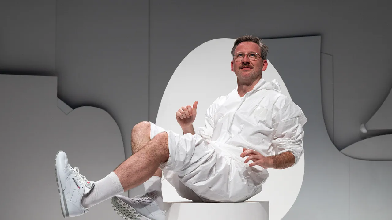 A performer wearing circular framed glasses and has a moustache, sits on a white plynth in a white jumpsuit, white socks and white sneakers. He has the pants of the jumpsuit pushed up, above his knees, and is leaning back with his legs bent infront of him. He is in front of a while egg shape, on a grey stage backdrop. He looks up, into the distance with a small smile on his face.