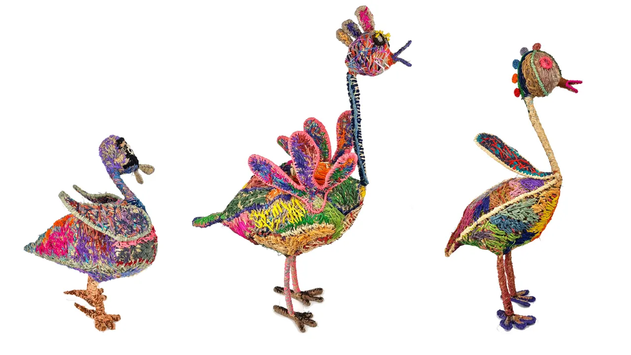 A photo of three sculpture pieces. The pieces are of birds, with long necks, made of brightly coloured straw in a stitched style. All three birds are in a line, all facing to the right..