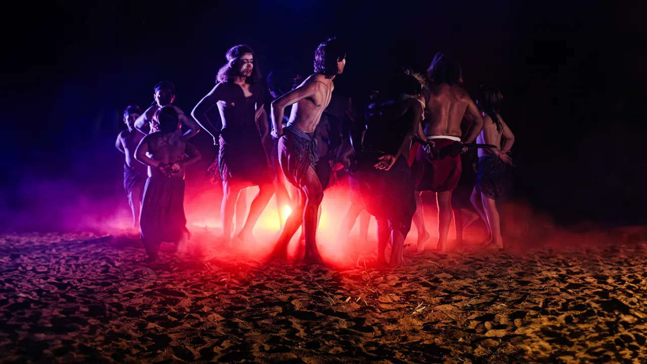 A photo of Indigenous dancers moving in a circle around a central red light. They are outdoors on sand under a dark sky.