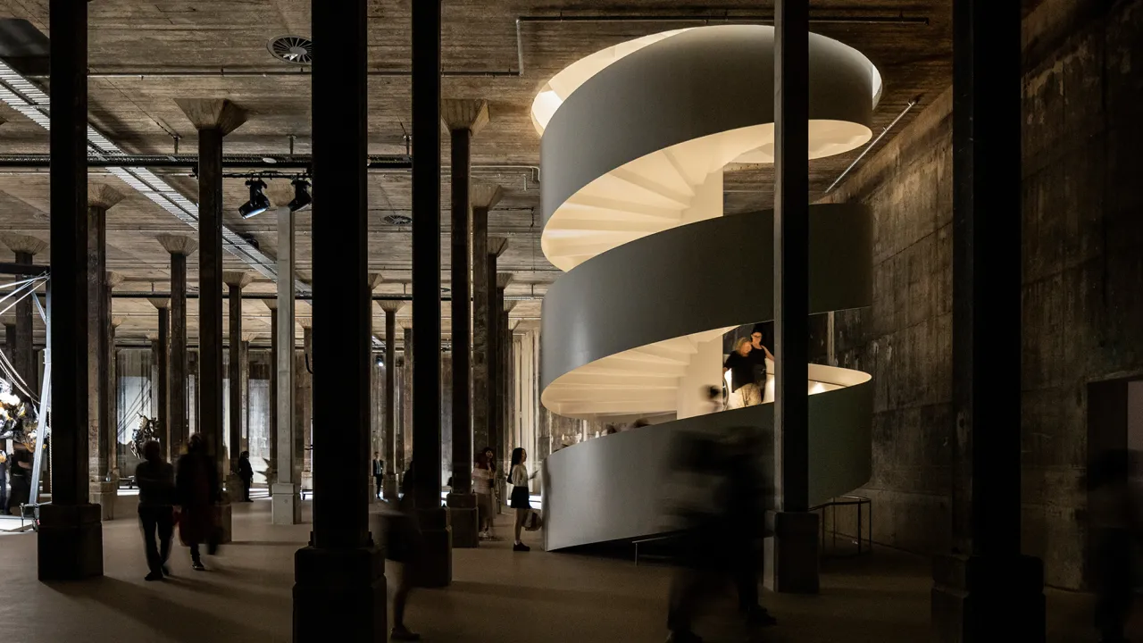 A photo of a glowing cylindrical stairwell positioned in a dark, industrial room. There are many mental pillars within the room and people walking up the stairs.
