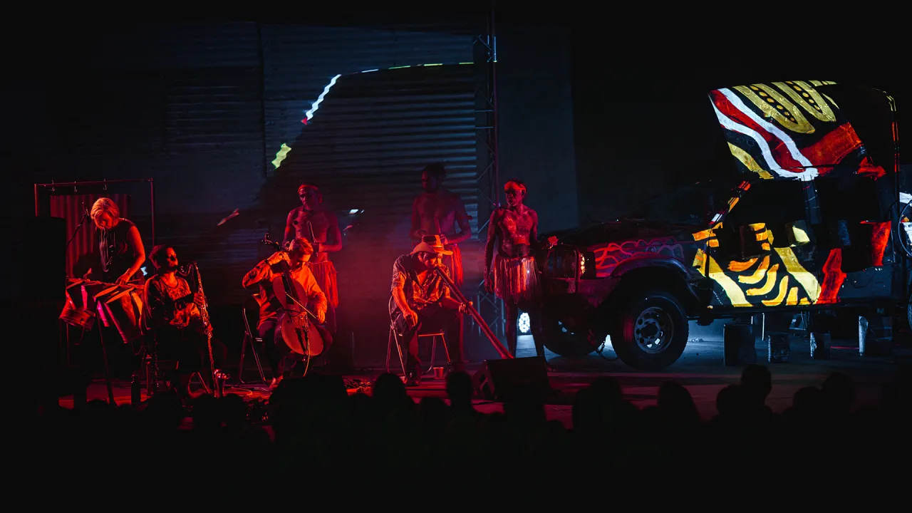 A photo of a band on a stage. The image is quite dark and moody, the musicians are glowing in a red light, in front of a semitruck that has an art work projected onto its side.