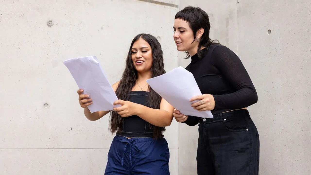 Two women holding scripts in a white room