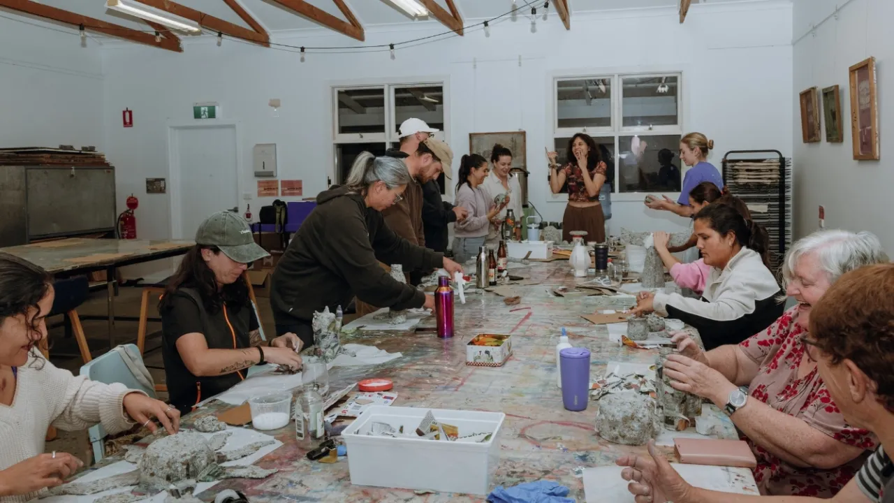Still image in landscape orientation showing adults gathered around long table with Paper Mache  workshop facilitator Cami laughing, in the Print Room at a Cannery Arts Centre workshop in Esperance,  Western Australia.