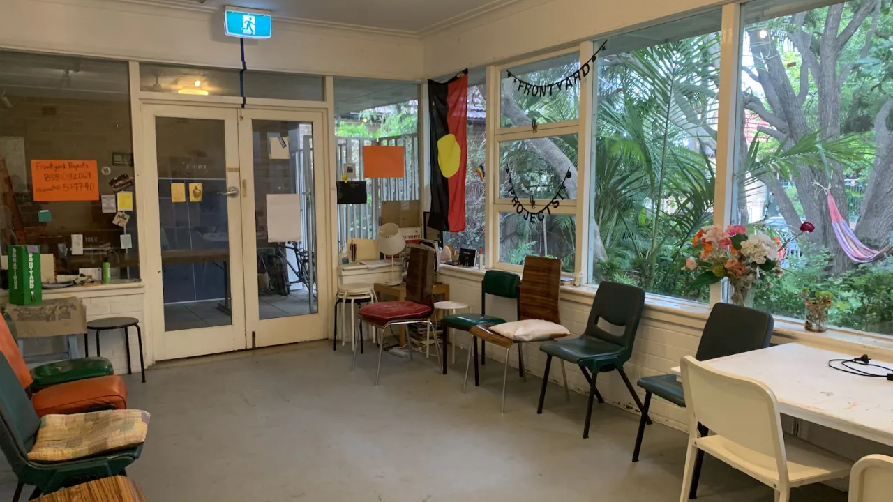 The Workshop Room at Frontyard features an empty space that you can make your own! An assortment of secondhand chairs line the walls and the windows facing onto the green garden. An Aboriginal Flag hangs from the ceiling in front of the windows along with a homemade banner reading, "Frontyard Projects." There are posters on the windows and the front door, which leads to our verandah. 