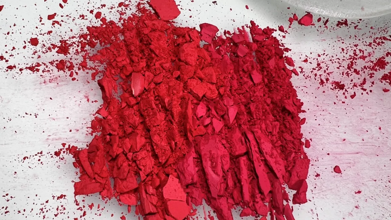 Close-up photograph of vivid red cosmetic pigment crushed into flakes and powder on a white surface. A large pile of fragmented red makeup sits in the centre, with fine pigment dust scattered around it. A translucent glass mulller is partially visible in the upper-right corner.