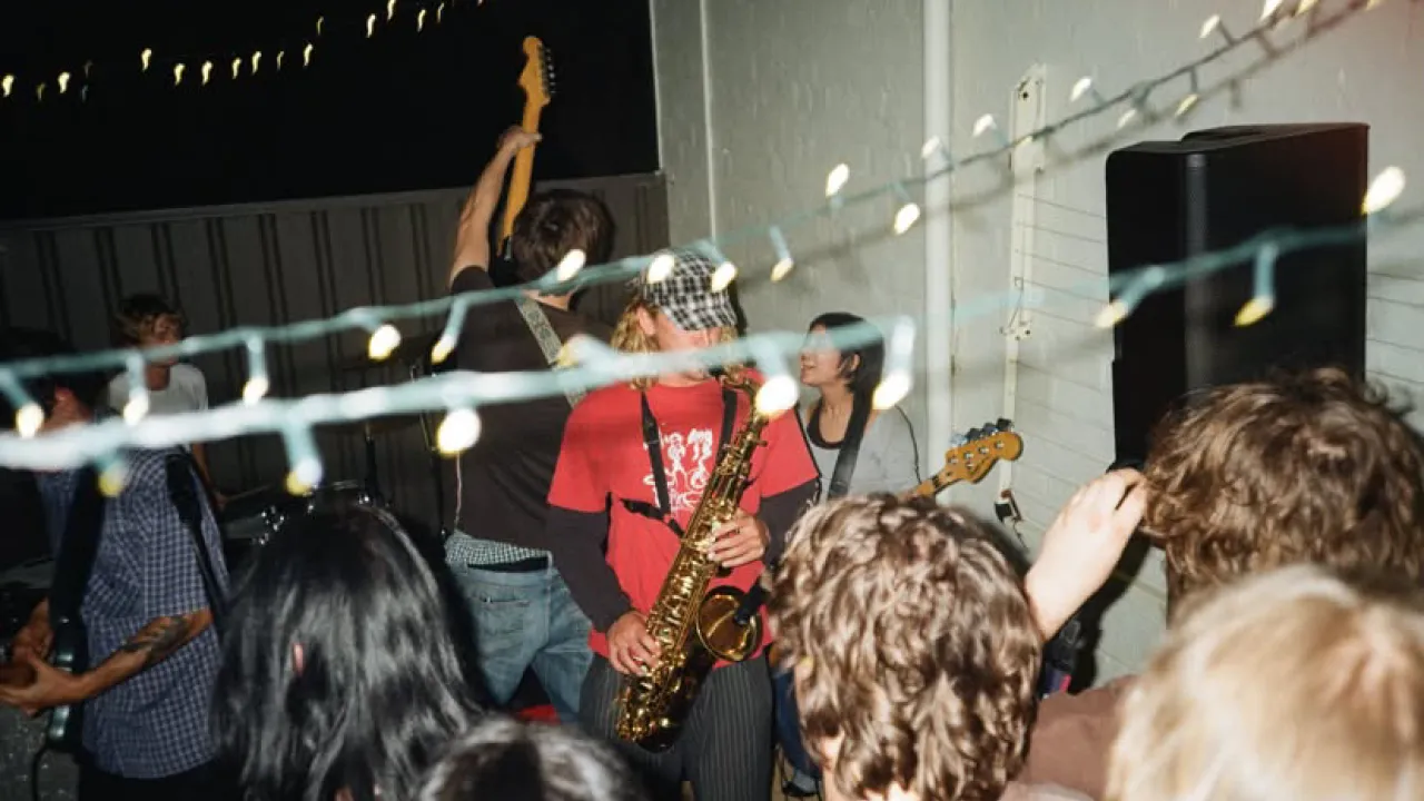 A musician in a red shirt playing saxophone with audience members watching in the foreground.