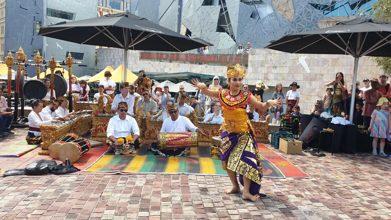 A Balinese dancer in an ornate gold, red and purple costume dances Tari Margapati in front of a Balinese gamelan percussion orchestra with 18 performers sitting behind various kinds of metallophones and gongs at Federation Square in Melbourne.