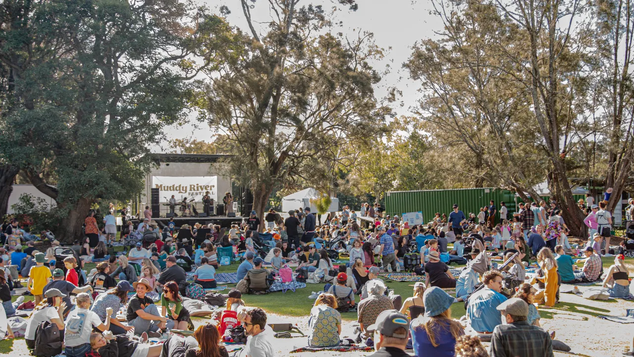 Families sitting on picnic blankets under trees with Muddy River Festival stage in the background