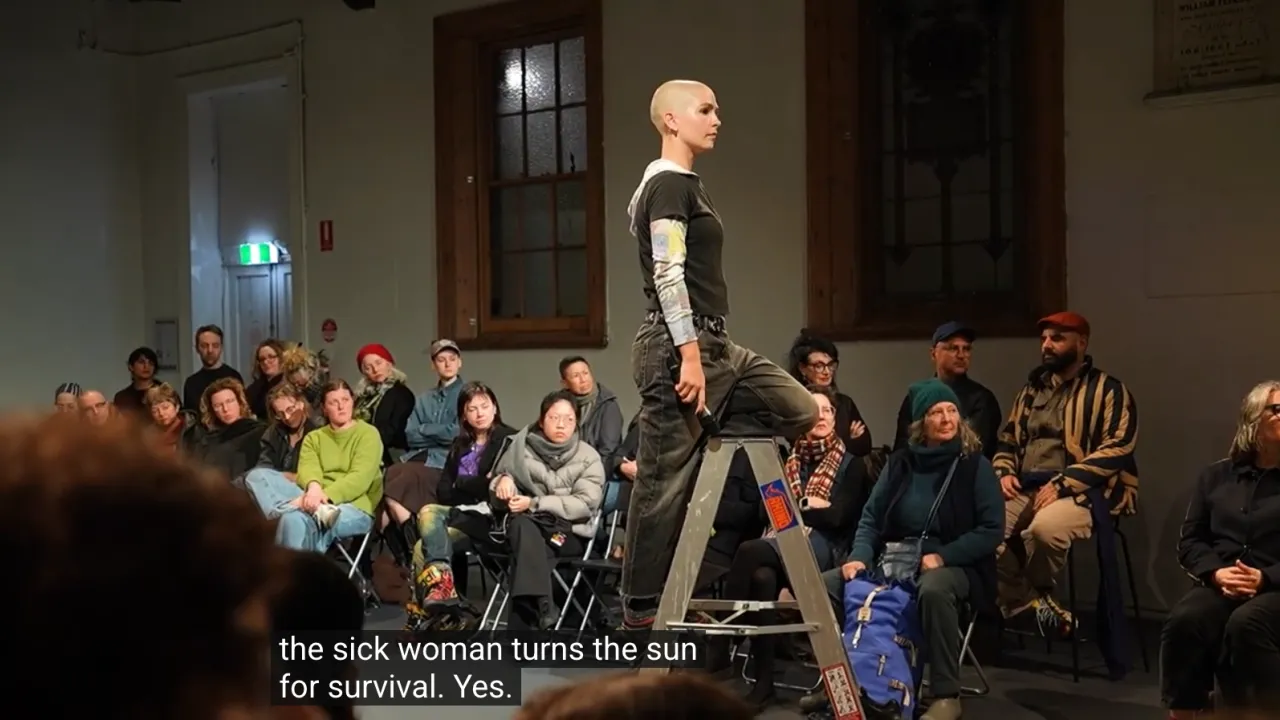 Image from showing of work in progress performance, ‘After Agatha’ by Amaara Raheem. The image features performer Gemma Sattler standing on a ladder, with an audience watching on. The image is a screen shot of video documentation captured by Anna White.