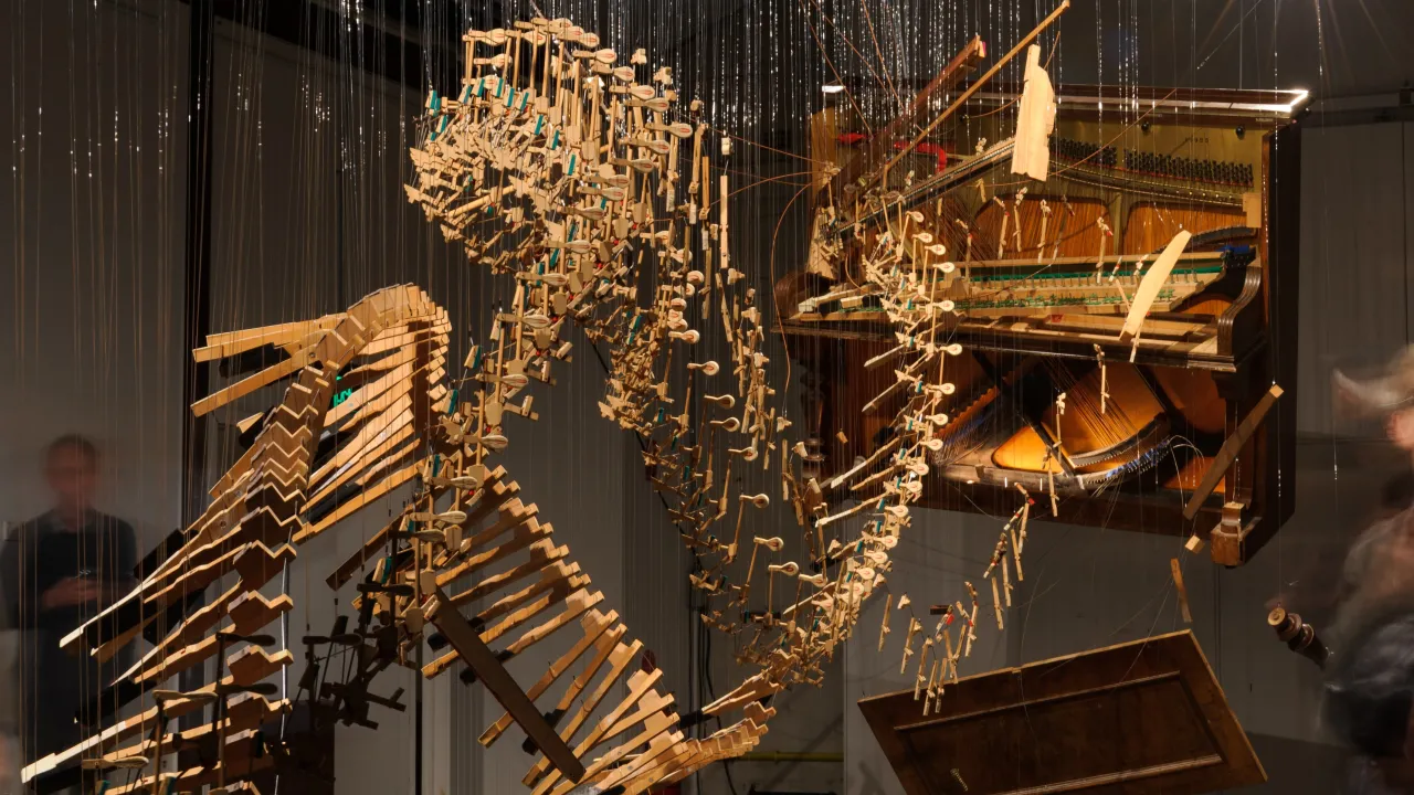 A deconstructed piano is suspended in a space with the body of the piano hanging separately from all the keys, shaped in a curved pattern. Blurred figures of people in the space look at the installation.
