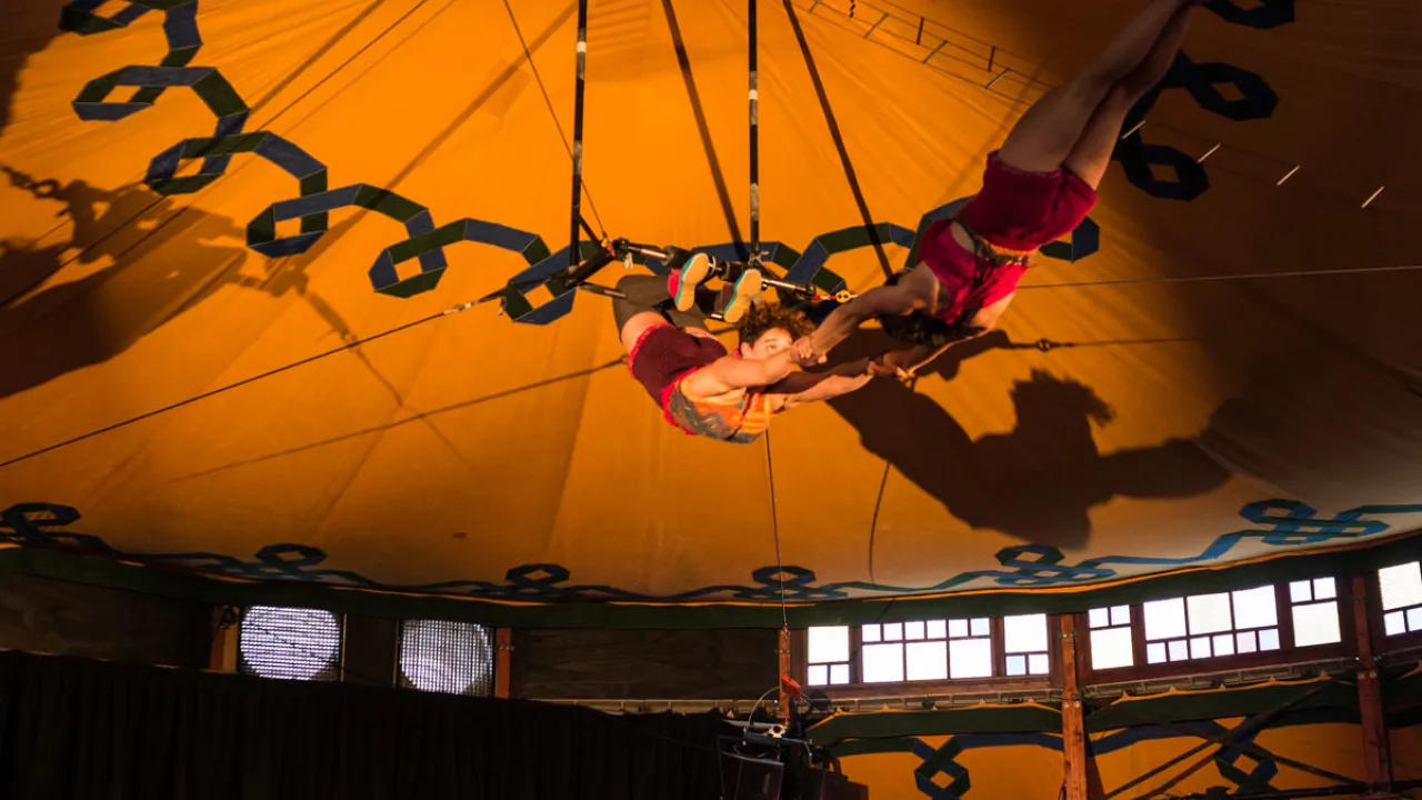 Two aerial artists flying against the yellow canopy of The Melba Spiegeltent.