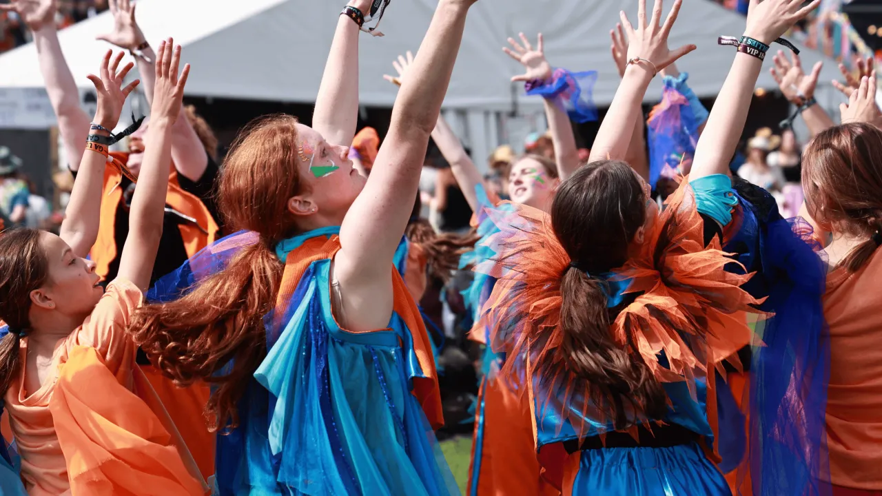 Group of dancers wearing bright blue and orange outfits standing in a circle with their arms outstretched above their heads at a large music festival