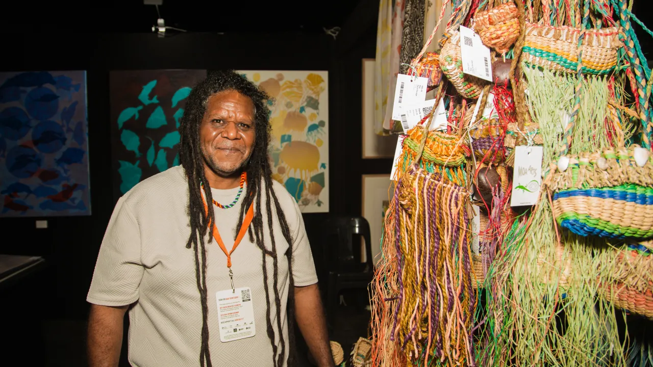 Solomon Booth standing in Moa Arts Booth at the Darwin Aboriginal Art Fair with screen prints and raffia bags in shot.