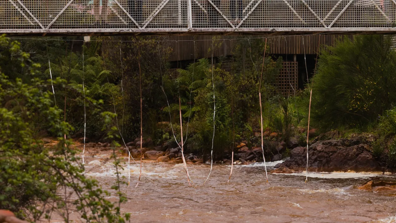 A pedestrian footbridge spanning a flowing river with copper plates dangling into the water.