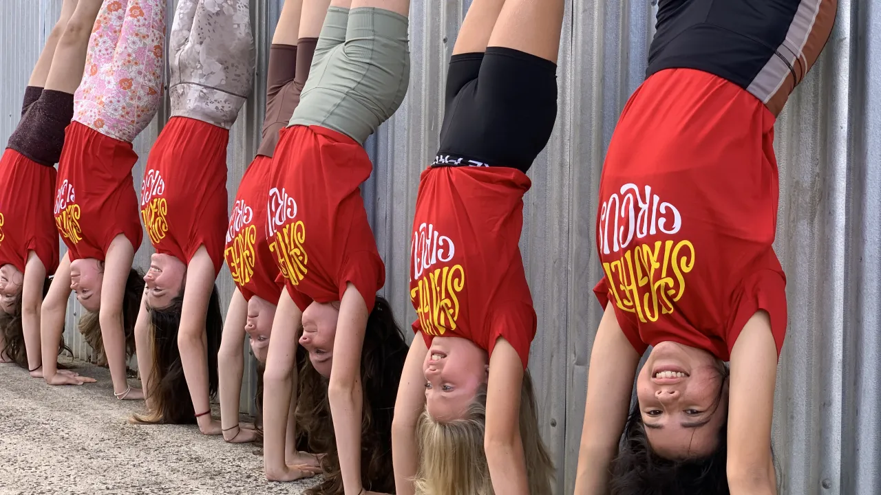 Six young circus performers do handstands in a row against a corrugated metal wall. They wear red Spaghetti Circus shirts and colourful shorts or leggings. The performer closest to the camera smiles while balancing upside down.