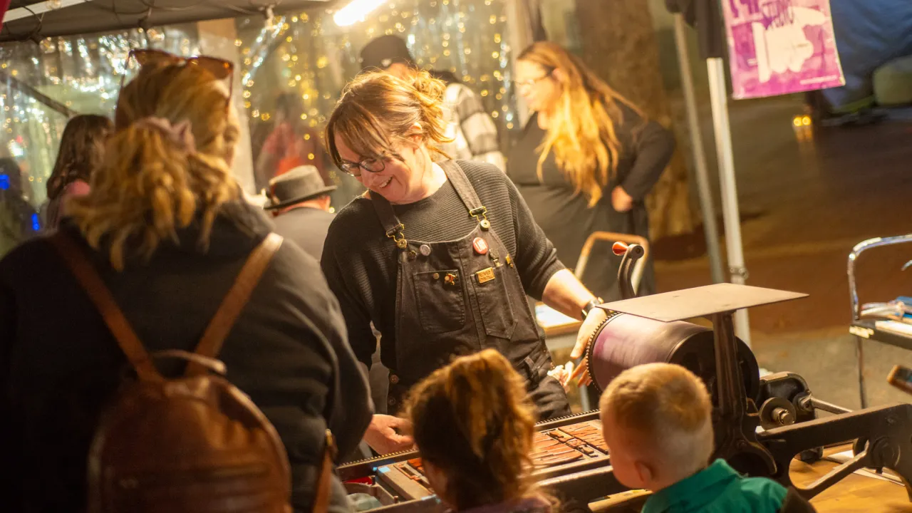 Kelly Lye of Hopscotch Press smiles as she demonstrates a vintage proof press at Moveable Type Studio's stall during A Winter's Night in Tamworth, 2026, while two young children watch the wooden type being inked. Fairy lights glow behind the marquee.