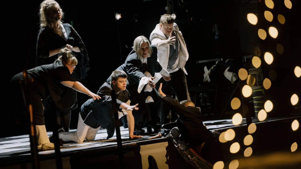 Members of Second Echo Ensemble perform together on a darkened stage, gathered around a performer off stage on a lower level. Several members reach to pull the performer up onto the stage, under warm theatrical lighting and out-of-focus lights glowing in the foreground.
