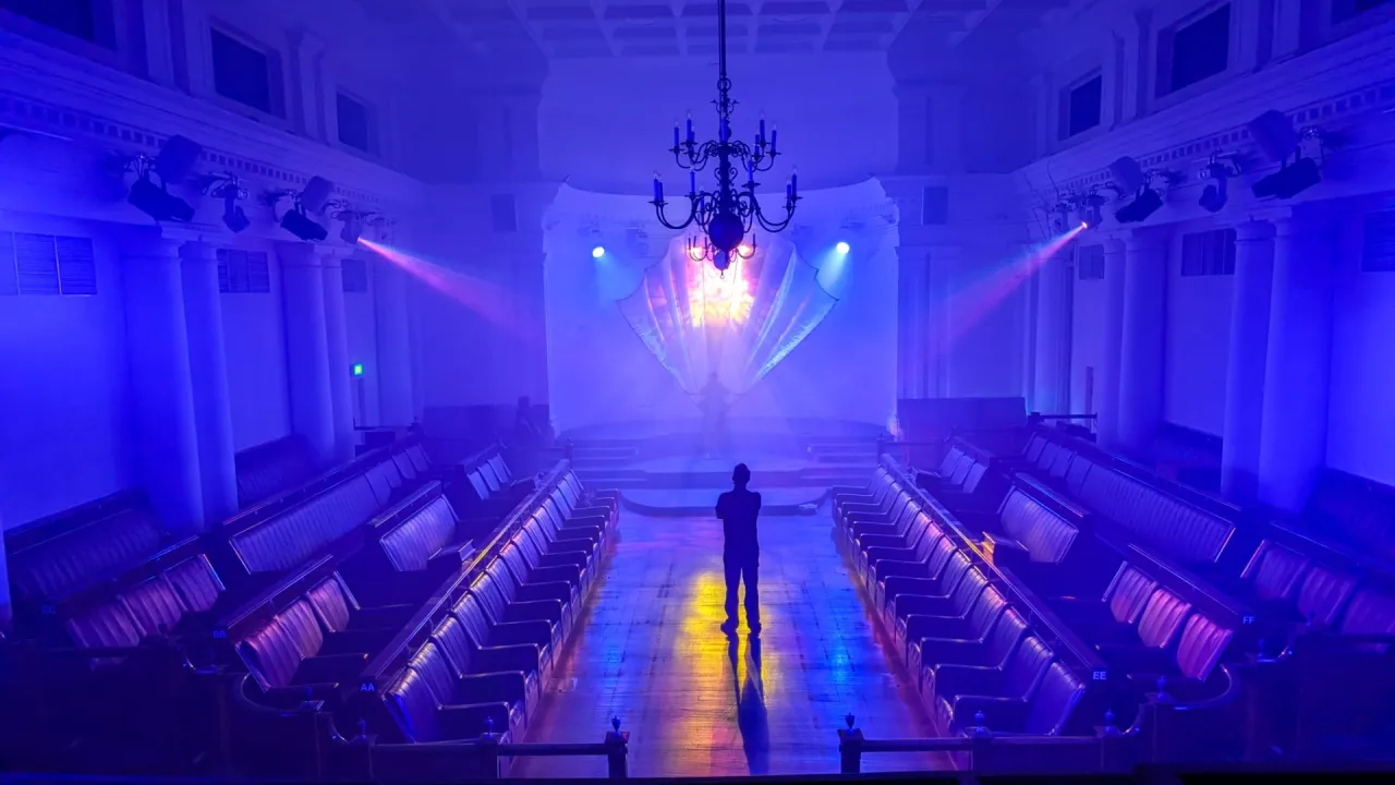 Theatre space illuminated in blue and purple light, with a lone figure standing in the centre aisle facing a glowing stage
