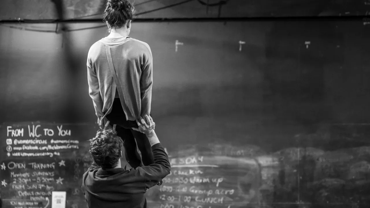 Black and white photograph taken inside a circus training space. Two people are mid-acrobatic lift — one person crouches low, gripping the hips of a second person who is raised up onto their shoulders, back to camera, wearing a cross-back top. Behind them a large chalkboard wall displays Women's Circus branding and a daily schedule including warm up, group acro, lunch, and performance. Black rigging infrastructure is visible overhead.
