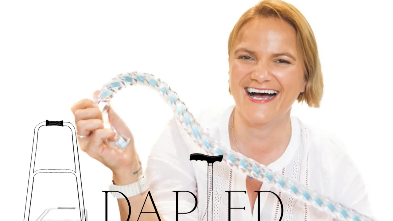 White background, woman with blond hair and a huge smile jauntily holding a walking stick with the stylised word Adapted written over the image. the A is a stylised A frame walker and the T is a stylised walking stick