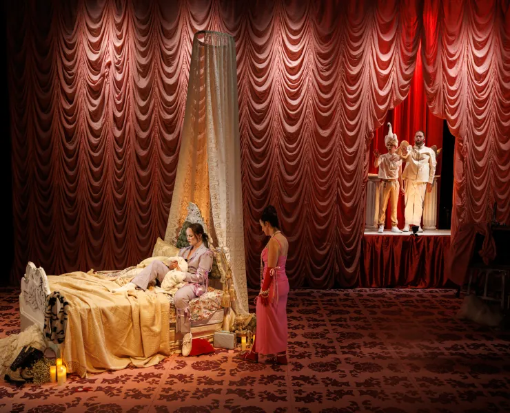 Stage screenshot of 2022 Iphis Production.  Opera by Elena Kats-Chernin and directed by Katy Maudlin.  Image features a wide stage screen shot with a pink curtain background.  A mother is arguing with her daughter, who is sitting on an elaborate bed. There are two god-like figures in the background of the photo.