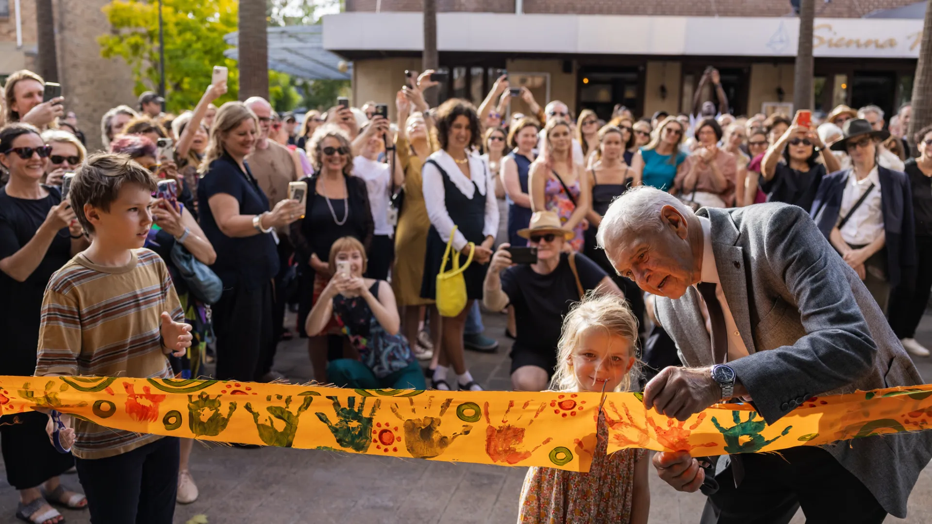 A photo of a large crowd applauding as an elderly man and a young girl cut a yellow ribbon that has a series of painted hands on it.