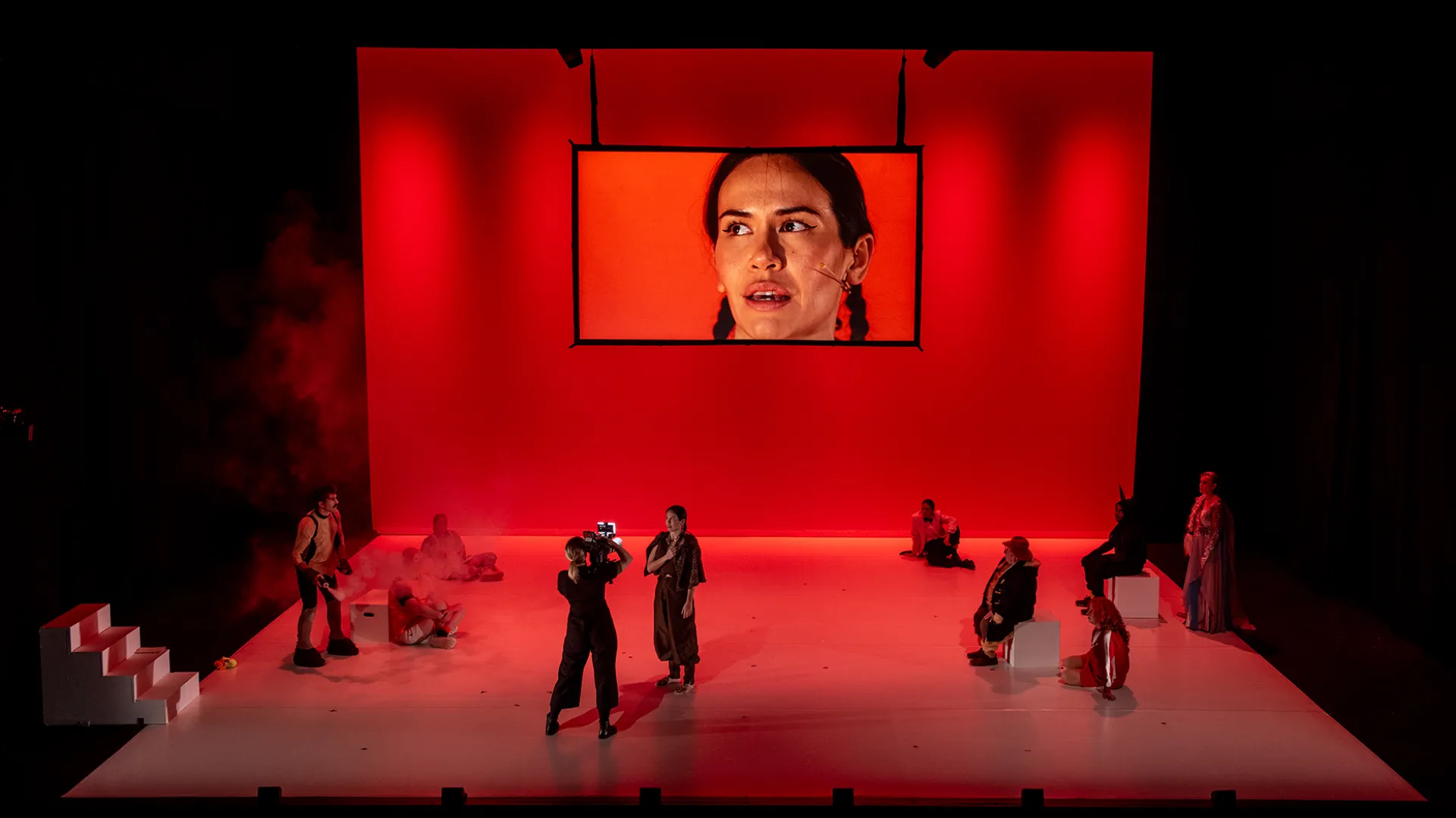 A photo of a performance on a large stage. The stage has a group of performers all dressed in black, spread across the stage. On the back of the stage is a screen that has a close up of a performers face. The stage is glowing red.
