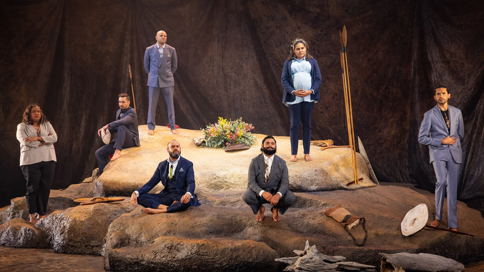 A staged photo of the theatre show 'The Visitors'. It shows seven indigenous performers standing and sitting no the set, made up of a large rock or cliff shape. The performers are all wearing corporate clothes but are barefoot. There are props of traditional spears, bags and tools. All the performers are staring out, past the camera into the distance.