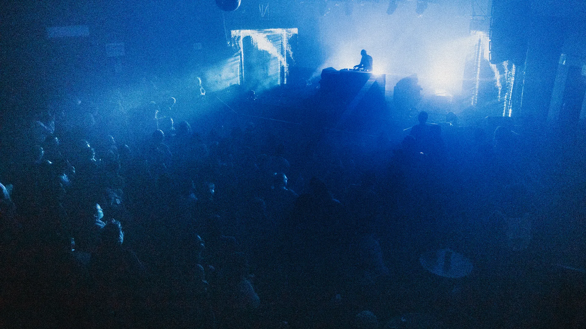 An edited photo of a night club. It is a photo from above and behind the crowd of a dark room, all watching a DJ standing behind the decks. There are bright white lights bursting behind the DJ, causing on their silhouette valuable. The room is dark and the shadows are deep blue. The image has been edited to be grainy and textured. 