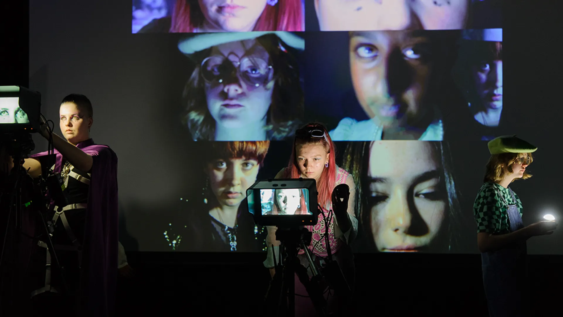 Three performers standing in front of a camera, with a collage of their faces projected on a screen behind them. They are teenagers in various makeup and costumes.