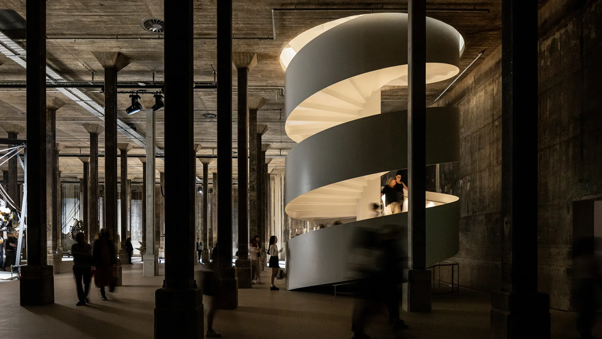 A photo of a glowing cylindrical stairwell positioned in a dark, industrial room. There are many mental pillars within the room and people walking up the stairs.