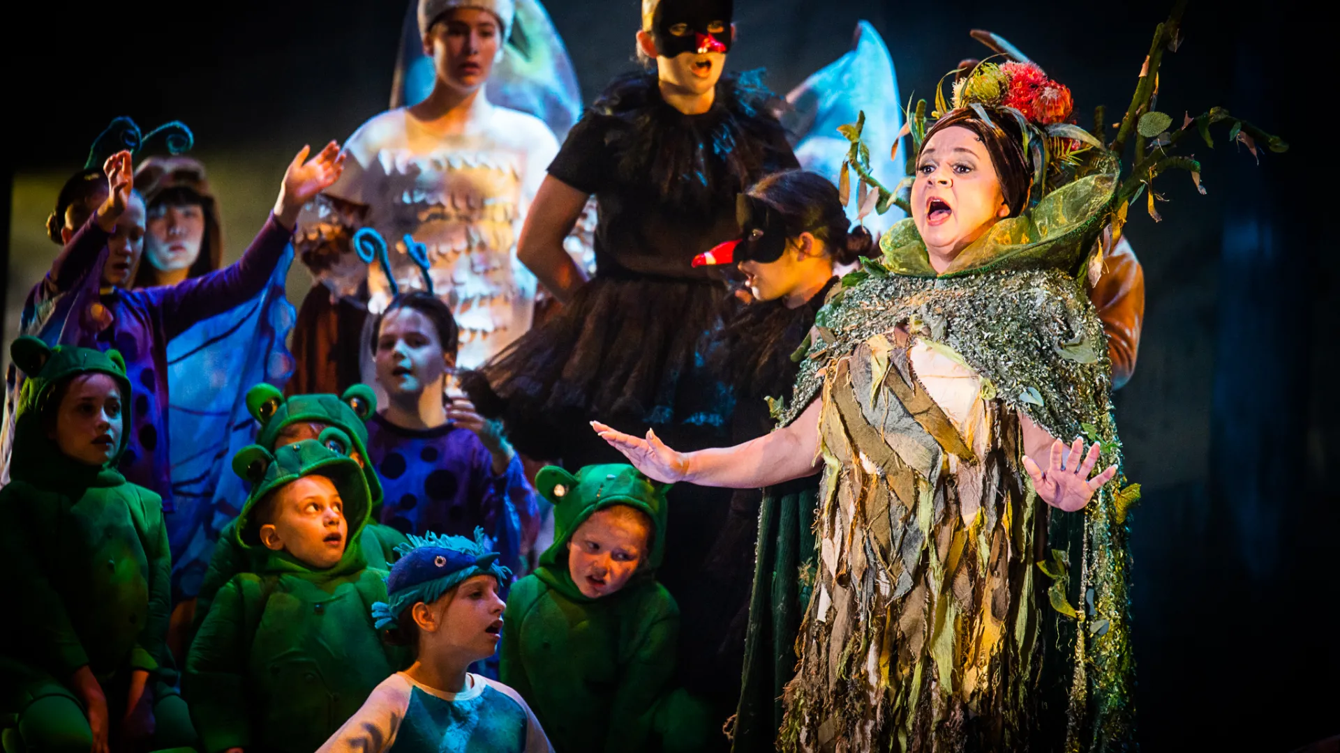 A photo of a group of performers all in elaborate and colourful costumes. There is one adult performer surrounded by a group of children performers. They are mid-song.
